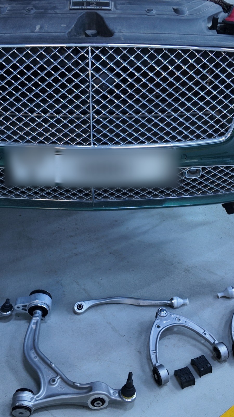 Front suspension control arms laid out under a green Bentley Continental GT during suspension work at Velokar