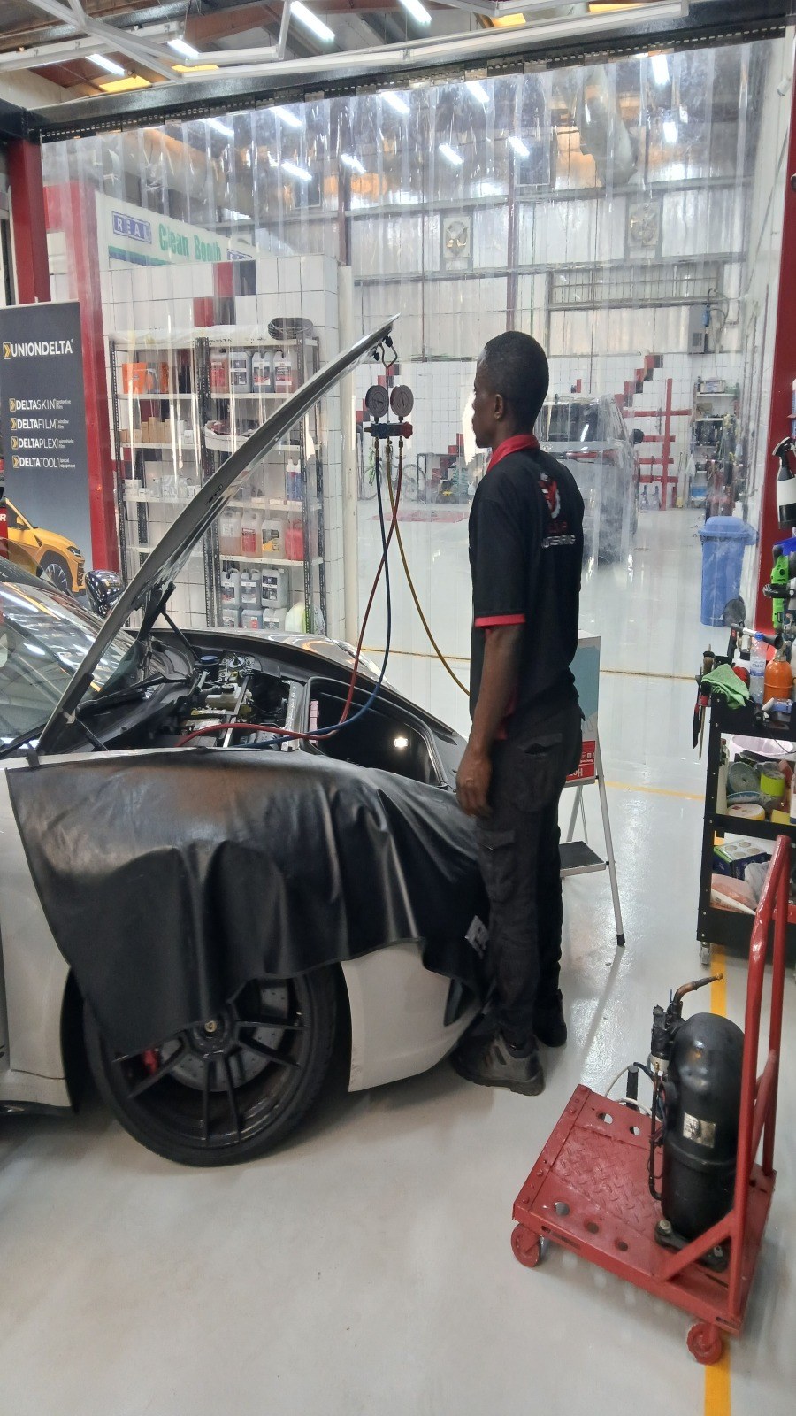 Velokar technician reading the AC manifold gauges connected to a white Porsche with the bonnet open, the recover-and-recharge station beside the car