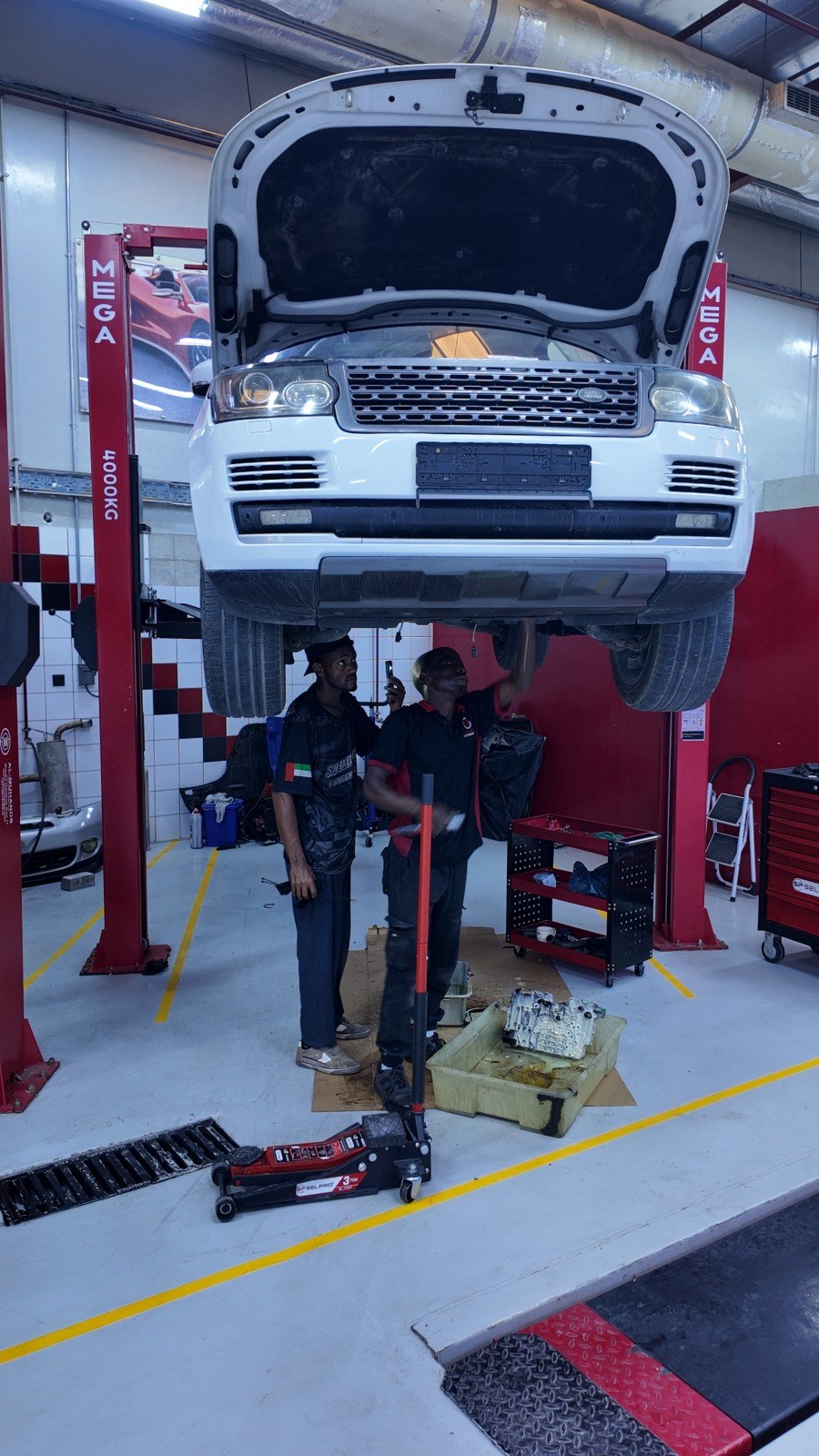 White Range Rover on the two-post lift at Velokar Auto Repairing, Al Quoz, two technicians underneath and the transmission valve body in a tray on the floor