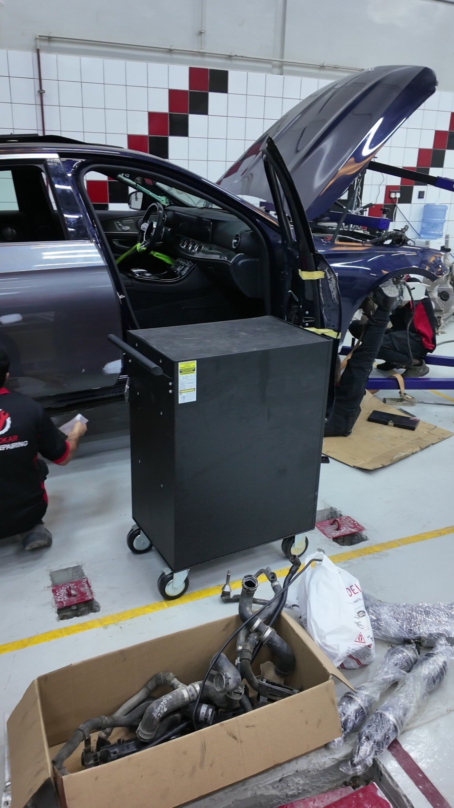 Cardboard box of removed hoses and wrapped new parts on the floor next to a navy sedan with its door open at Velokar