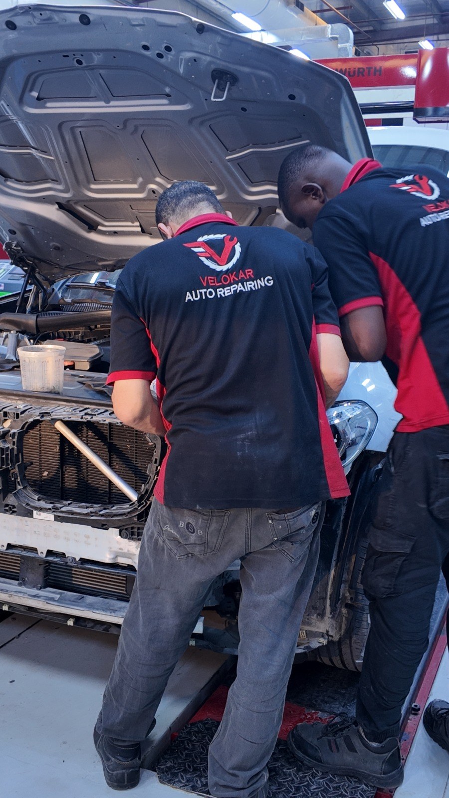Two Velokar technicians working at the front of a white car with the bonnet up and the front end stripped for access