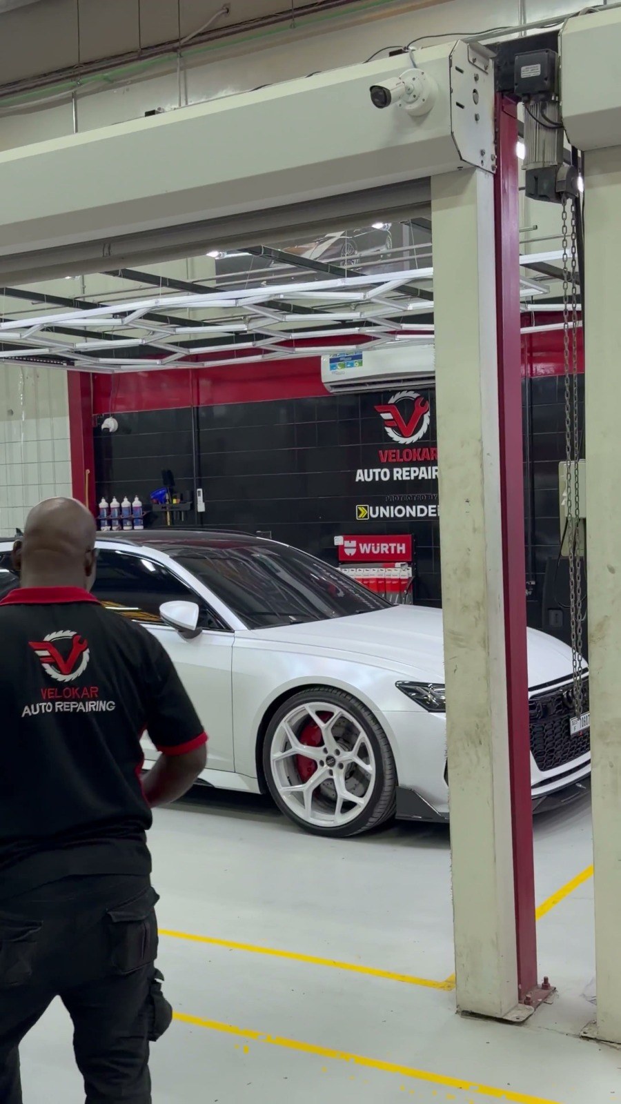 Velokar technician at a lift column in the Al Quoz workshop, a white car with red brake calipers in the bay behind him and the Velokar sign on the wall